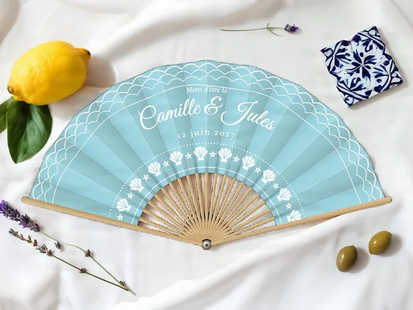 Éventail de mariage personnalisé « Bord de mer », vagues en bordure et arc de coquillages sous les prénoms, toile : bleu ciel, impression : blanc, posé sur une table de mariage