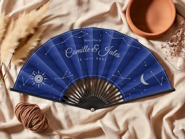 Éventail de mariage personnalisé « Céleste », constellations, soleil et croissant de lune semés autour des prénoms, toile : bleu marine, impression : argent, posé sur une table de mariage