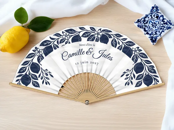 Éventail de mariage personnalisé « Citrons d'Amalfi », branches de citrons et fleurs encadrant les prénoms, toile : blanc, impression : bleu marine, posé sur une table de mariage