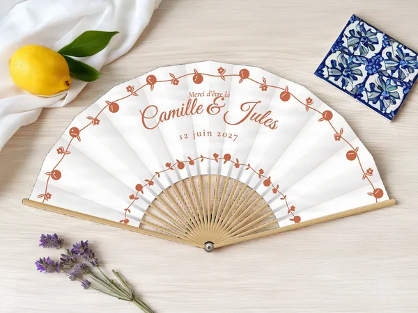 Éventail de mariage personnalisé « Dolce vita », guirlandes de petits agrumes et fleurs en arcs autour des prénoms, toile : blanc, impression : terracotta, posé sur une table de mariage