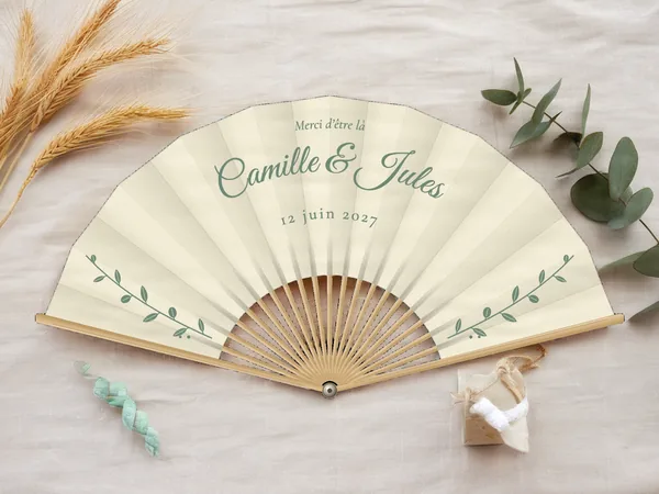 Éventail de mariage personnalisé « Eucalyptus », fines branches feuillues de part et d'autre sous les prénoms, toile : ivoire, impression : vert sauge, posé sur une table de mariage