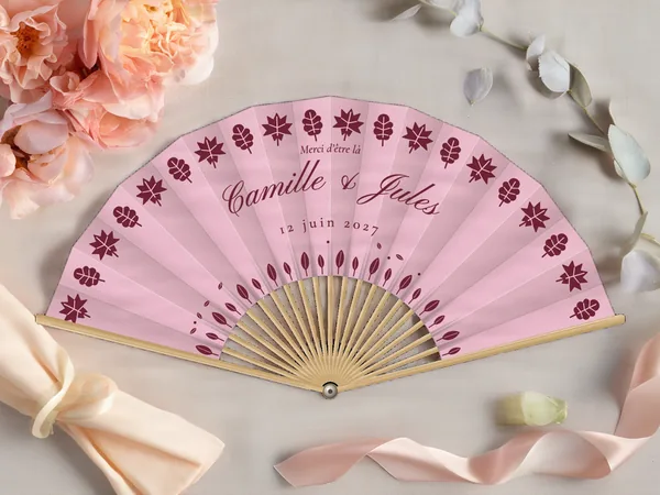 Éventail de mariage personnalisé « Feuilles d'automne », feuilles stylisées en frise sur tout le pourtour des prénoms, toile : rose poudré, impression : bordeaux, posé sur une table de mariage