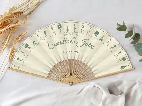 Éventail de mariage personnalisé « Fleurs des champs », petites fleurs des champs alignées en couronne autour des prénoms, toile : ivoire, impression : vert sauge, posé sur une table de mariage