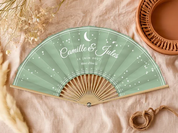 Éventail de mariage personnalisé « Lune et étoiles », croissant de lune et étoiles semées autour des prénoms, toile : vert sauge, impression : blanc, posé sur une table de mariage