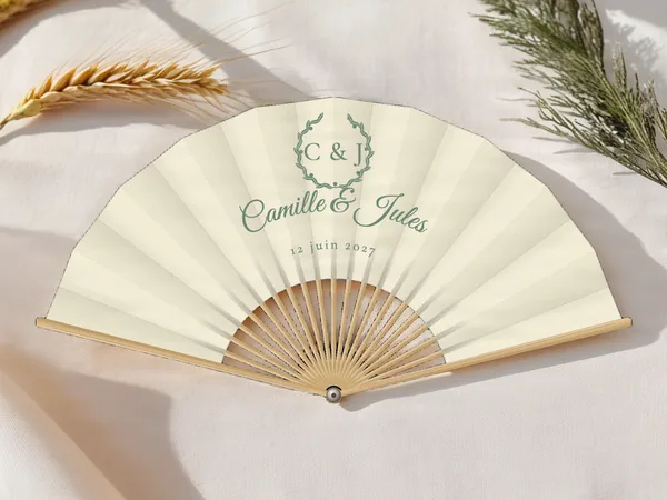 Éventail de mariage personnalisé « Couronne d'olivier », couronne de feuillage entourant les initiales au-dessus des prénoms, toile : ivoire, impression : vert sauge, posé sur une table de mariage