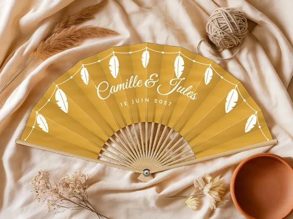 Éventail de mariage personnalisé « Plumes », plumes suspendues à une guirlande perlée le long du bord, toile : moutarde, impression : blanc, posé sur une table de mariage