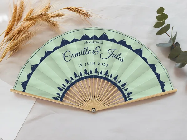 Éventail de mariage personnalisé « Sapins et montagnes », crêtes de montagnes en bordure et rangée de sapins en bas, toile : vert d'eau, impression : bleu marine, posé sur une table de mariage
